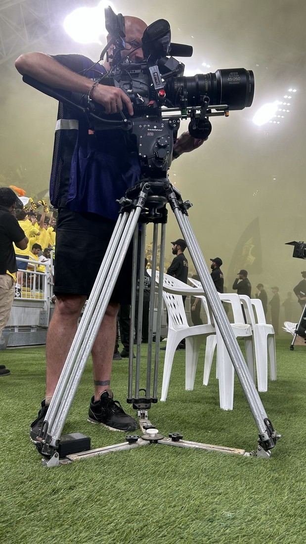 Netflix Saudi Pro League documentary, Jeddah: a camera operator on a cinema camera at pitch level, with yellow smoke from the stands behind